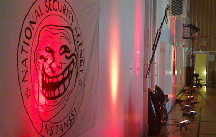 Instanssin valoja yhdeltä seinustalta kuvituskuva Instanssi 2014 -tapahtumasta. Seinällä National Security Agency Instanssi -logo, jonka keskellä on trollface. Valot osoittavat seinälle punaisina ja keltaisina